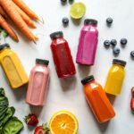 colorful bottles with smoothies beside carrots ginger leaves and berries